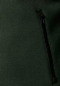 Groene ribbelstof met een gestructureerde uitstraling, voorzien van een zijzak met rits in zwart. De samenstelling is zacht met een aansluitend ontwerp.