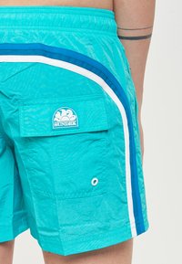 Pantaloni da bagno turchesi, materiale leggero, con strisce bianche e blu, tasca sul retro con patta e un'etichetta logo turchese.