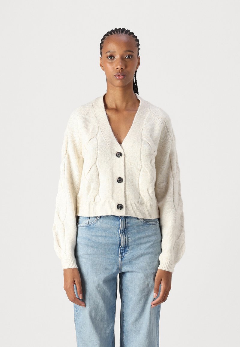 Cardigã creme de malha trançada com decote em V, fechamento com botões e mangas balão, combinado com jeans de cintura alta em azul claro.