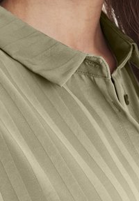 Oliwkowa bluzka o prążkowanej fakturze, z kołnierzykiem i detalem w postaci guzika, prezentująca delikatny wzór w pionowe paski.
