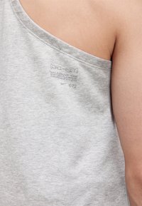 Gros plan sur une épaule portant un débardeur Nike Dri-FIT gris clair avec un petit logo embossé et un texte dans le haut du dos.