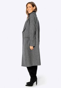 Cappotto lungo grigio in lana con reverse oversize, tasche laterali e silhouette dritta. Indossato con stivali neri al ginocchio.