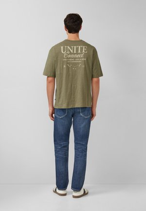 Man die met zijn rug naar de camera staat, draagt een olijfgroen T-shirt met de tekst "UNITE Connect" en een blauwe spijkerbroek, tegen een effen grijze achtergrond.