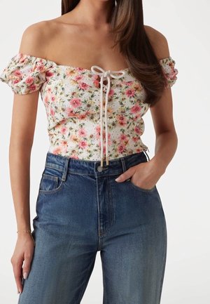 Woman wearing off-shoulder floral blouse with puff sleeves and high-waisted blue jeans, one hand in pocket against white background.