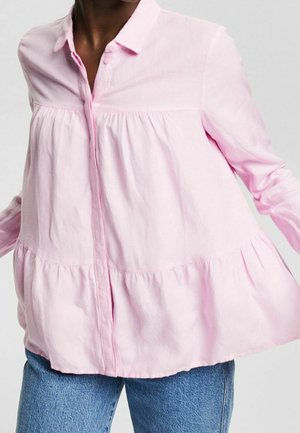 Femme portant un chemisier ample rose pâle à manches longues avec des coutures à volants, assorti à un jean bleu en denim, sur un fond uni.