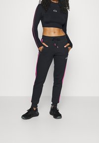 Musta lyhythihainen crop-top, jossa on pieni logo ja vaaleanpunaiset sivu raidat; yhteensopivat mustat jogging-housut vaaleanpunaisilla yksityiskohdilla ja sivutaskuilla.