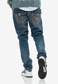 Persona vistiendo jeans azules con bolsillos traseros cosidos, zapatillas blancas que muestran "AMIRI" en la suela y una sudadera verde, caminando de espaldas.