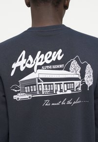 Donkergrijze sweatshirt met witte grafische print van een hut en bergen, tekst "Aspen ALPINE RESORT," en een autodesign.