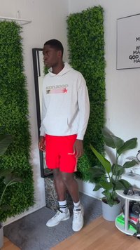 Hoodie cinza de manga longa com logotipo, calções vermelhos e ténis brancos. A cena apresenta vegetação e um fundo espelhado com plantas de interior.
