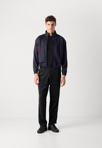 Ναυτική αδιάβροχη ζακέτα με κόκκινες λεπτομέρειες, ελαστικούς μαν sleeves, και φερμουάρ μπροστά, σε συνδυασμό με μαύρο παντελόνι και μαύρα αθλητικά παπούτσια. Λείος υφασμάτινος ιστός.