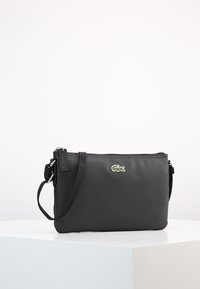 Borsa a tracolla sintetica nera con superficie testurizzata, chiusura con zip e tracolla regolabile. Presenta un accento del logo verde di Lacoste.