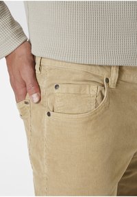 Beige Corduroy-Hosen haben eine schmale Passform, Seitentaschen und kontrastierende Metallbeschläge mit einer gerippten Stoffstruktur.