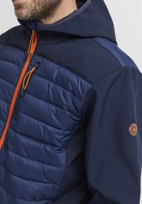 Mørkeblå jakke med polstret øverste del, oransje glidelås og hette. Har en glatt tekstur med detaljert logo på ermene.