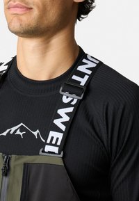 Zwart langemouwen shirt met ribbeltextuur, voorzien van een grafiek van bergen; verstelbare banden met witte letters; grijs en olijfgroen overall.