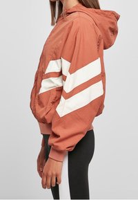 Rostfärgad bombarjacka med vita chevronränder. Tillverkad av lätt material, har en huva och elastiska ärmslut.