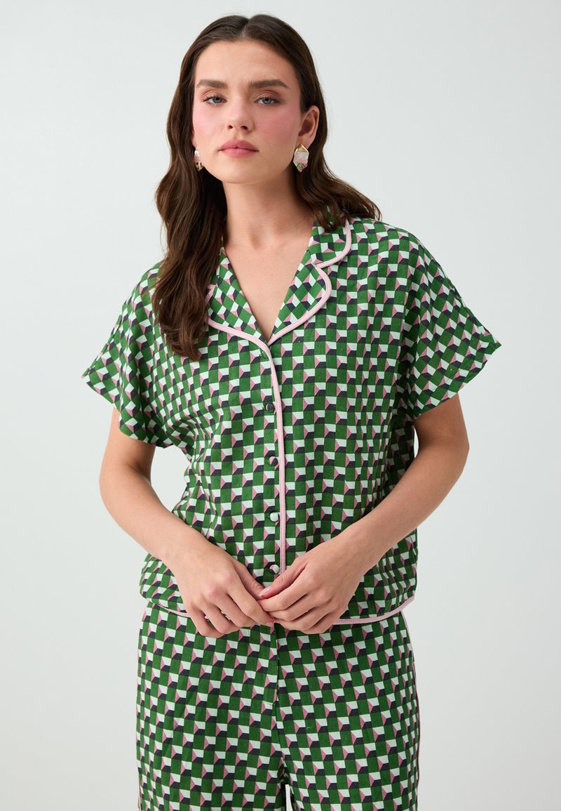 Femme aux longs cheveux foncés portant une chemise boutonnée à manches courtes à motif géométrique vert et blanc et un pantalon assorti, debout devant un fond uni.