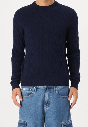 Homme portant un pull à torsades bleu marine et un pantalon cargo en jean bleu clair, debout les mains détendues le long du corps.