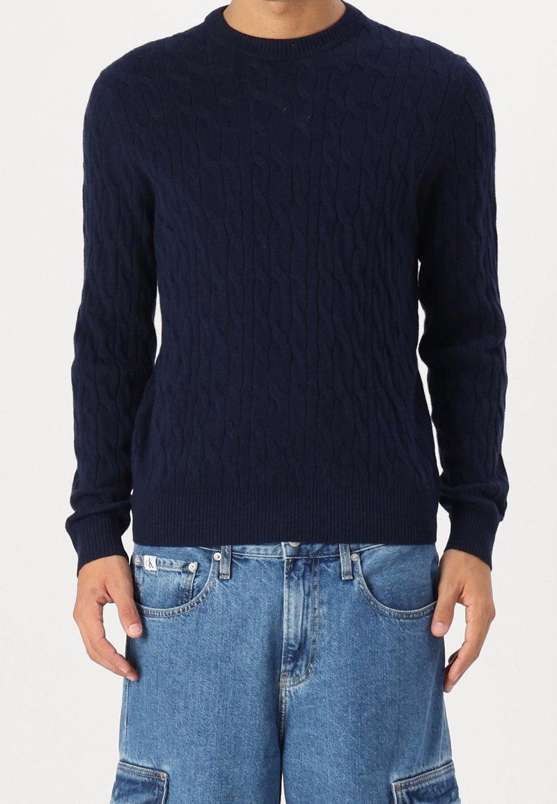 Homme portant un pull à torsades bleu marine et un pantalon cargo en jean bleu clair, debout les mains détendues le long du corps.