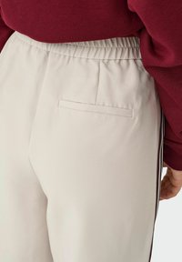 Jasnobeżowe dresy z elastycznym paskiem w talii, wyposażone w jedną kieszeń z tyłu oraz kontrastowe paski po bokach. Gładka tekstura, casualowy design.