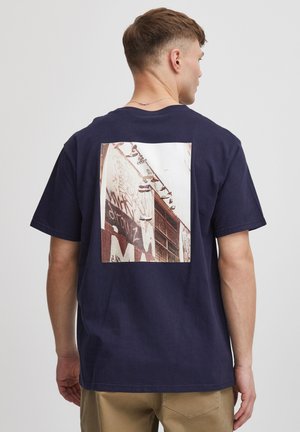 Marineblaue Baumwoll-T-Shirt mit einem rechteckigen Grafikdesign auf der Rückseite, das eine mit Graffiti bedeckte Wand mit Schuhen zeigt. Kurze Ärmel und Rundhalsausschnitt.