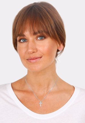 Silver pendant necklace featuring a small cross, set on a thin chain. The wearer has a light-toned shirt and smooth, highlighted skin.