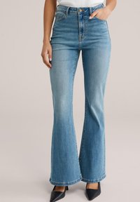 Jeans evazați albastru deschis cu talie înaltă din denim cu decolorare subtilă, cinci buzunare și închidere cu nasture, asortați cu pantofi negri cu vârf ascuțit.