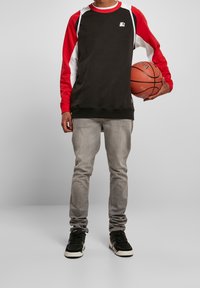 Persona de pie sosteniendo un balón de baloncesto, vistiendo una camiseta de manga larga negra y roja, jeans grises y zapatillas negras, contra un fondo liso.