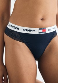 Marineblauwe bikini-slips met kanten zijaccenten en een witte elastische tailleband met "TOMMY HILFIGER"-merknaam en logo.