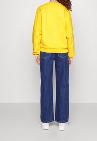 Sudadera amarilla de corte holgado, con puños y dobladillo de canalé, combinada con jeans de pierna ancha en azul oscuro que cuentan con bolsillos traseros y costuras visibles.
