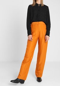 Schwarzes Langarmhemd mit weißen Polka-Dots, kombiniert mit orangefarbenen, plissierten Hosen. Das Modell trägt schwarze Stiefeletten.