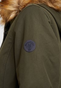 Abrigo verde oliva con capucha forrada de piel, dobladillo redondeado y un parche de logo azul marino en la manga izquierda con texto en relieve.