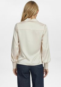 Blusa in crema di raso con colletto, maniche voluminose e polsini arricciati. Presenta una schiena pulita con un sottile design a yoke. Indossata con jeans scuri.