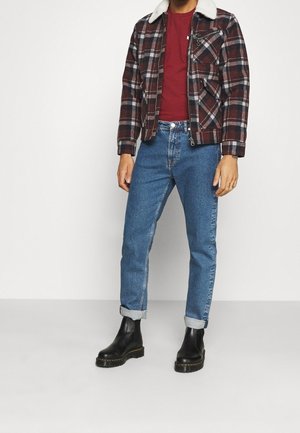 Mies, jolla on punainen paita, ruskea ja musta ruudullinen takki valkoisella kauluksella, siniset rullatut farkut ja mustat nahkasaappaat, seisomassa valkoisella lattialla.