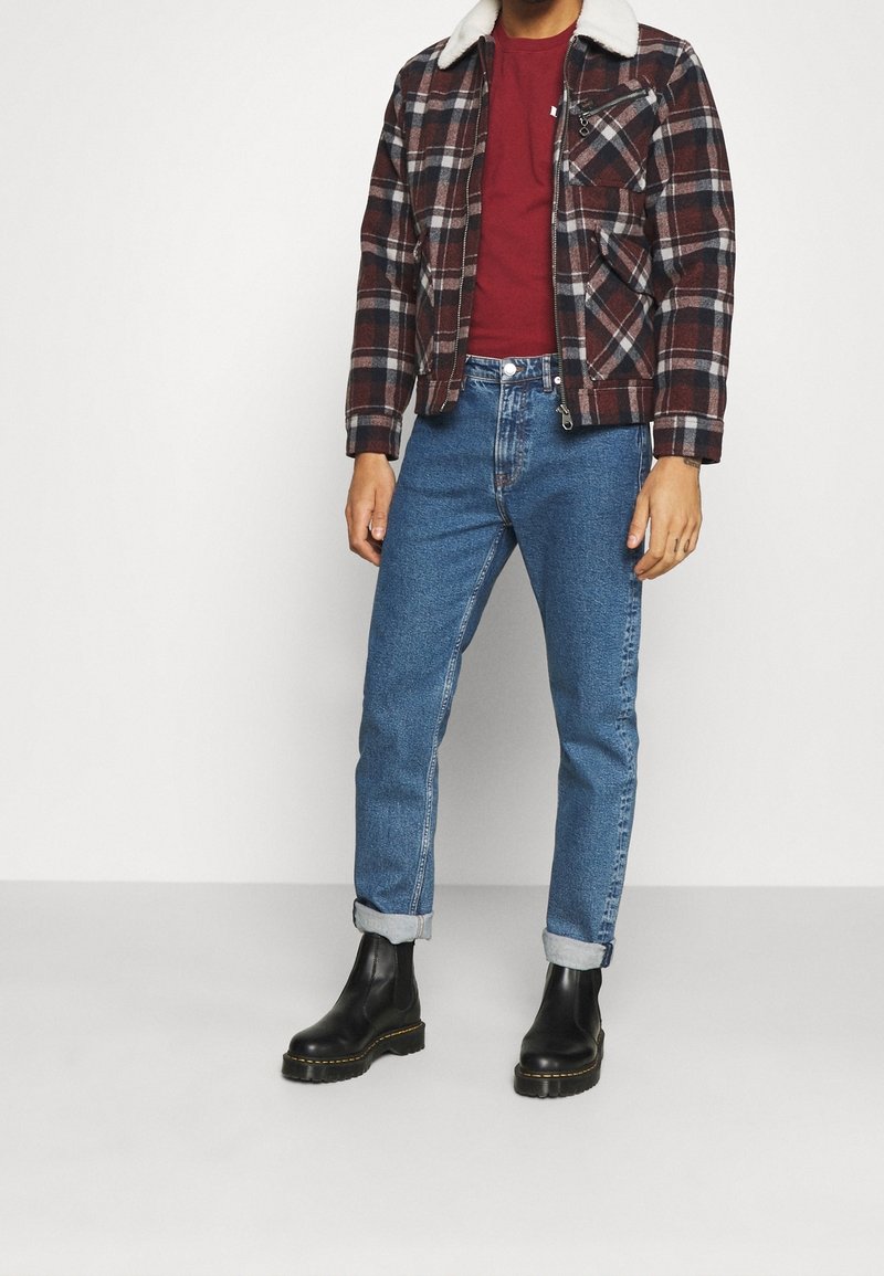 Homme portant une chemise rouge, une veste à carreaux marron et noire avec un col blanc, un jean bleu retroussé et des bottes en cuir noir, debout sur un sol blanc.
