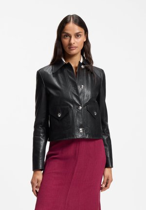 Frau trägt eine schwarze Lederjacke mit silbernen Knöpfen und Vordertaschen über einem hoch taillierten, dunkelrosa Rock und steht vor einem schlichten weißen Hintergrund.