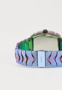 Silberne und lila metallische Uhr mit einem strukturierten Armband mit wellenförmigen Mustern und einem quadratischen Zifferblatt. Markenlogo sichtbar an der Schließe.
