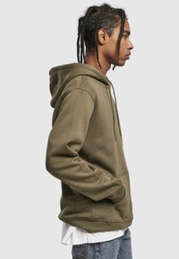 Bluza z kapturem w kolorze oliwkowej zieleni, wykonana z miękkiego materiału. Posiada przednią kieszeń i sznurek do regulacji kaptura, noszona na białej koszulce z niebieskimi dżinsami.