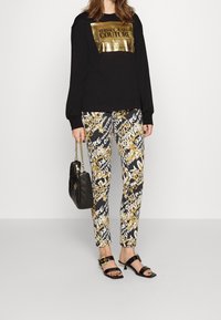 Sudadera negra de manga larga con logotipo dorado, combinada con pantalones de diseño en blanco y negro con acentos dorados. Bolso negro y sandalias.