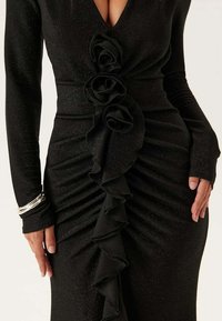 Černé přiléhavé šaty z lesklé látky. Mají hluboký výstřih do V, dlouhé rukávy a volánkový design s růžovými akcenty vpředu.