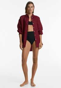 Burgundi vászon ing, felhajtott ujjakkal, gombos elöl és egyetlen mellzsebbel, fekete bikini top és alsó fölött viselve.