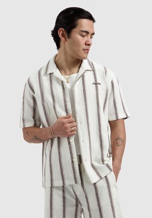 Man draagt een wit en beige verticaal gestreept kort mouwhemd en bijpassende korte broek, gouden sieraden en tatoeages op beide onderarmen.