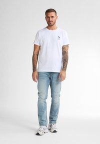 T-shirt bianca a girocollo con una piccola grafica colorata sul lato sinistro del petto, abbinata a jeans azzurri strappati e scarpe da ginnastica bianche.