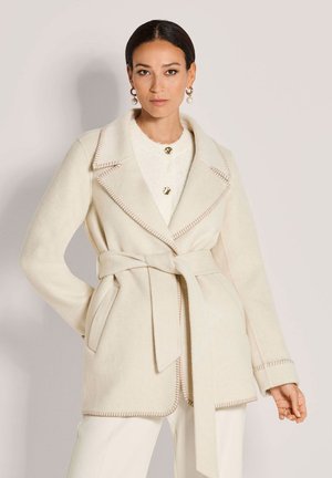 Frau mit dunklen, zurückgebundenen Haaren trägt einen cremefarbenen, taillierten Mantel mit großen Revers und Steppnähten über einem weißen Outfit sowie goldene Perlenohrringe.