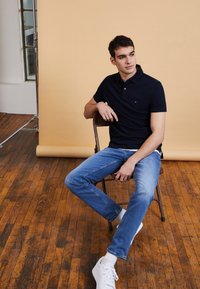 Dunkelblaues Polo-Shirt mit kleinem Logo, hellblaue Jeans und weiße Sneakers. Modell sitzt auf einem Holzstuhl vor einem beigen Hintergrund.