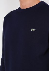 Sweatshirt en coton bleu marine avec un col rond, arborant un petit logo crocodile brodé vert et blanc sur la poitrine.