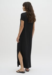 Black maxi dress with short sleeves, side slit, and a relaxed fit, made from a smooth fabric. Worn with black flip-flops.