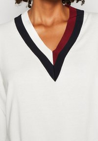 Weißer Pullover mit V-Ausschnitt und einem schwarz-bordeaux gestreiften Rand. Weiche Textur, lockere Passform, ohne Muster oder sichtbare zusätzliche Verzierungen.
