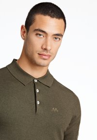 Olivgrön polo med texturerad stickning, tre-knaps placket och broderad logotyp på bröstet. Har korta ärmar.