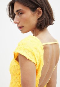 Robe en dentelle jaune avec broderie florale, manches courtes et design à dos ouvert. Le tissu est texturé, ce qui renforce l'intérêt visuel.