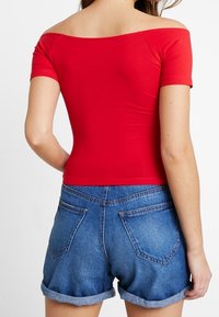 Kvinde iført en rød off-shoulder top og oprullede blå denimshorts, vist fra bagsiden mod en ensfarvet hvid baggrund.